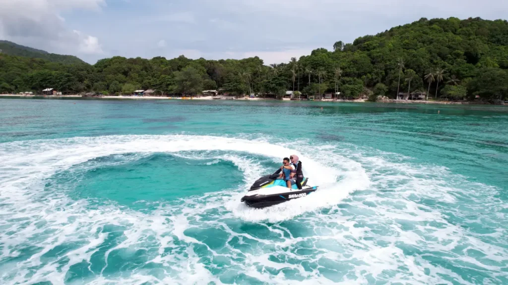 Dua orang menikmati jet ski di perairan jernih Karimunjawa, menciptakan ombak putih. Pemandangan pulau hijau di latar belakang, cocok untuk paket wisata air