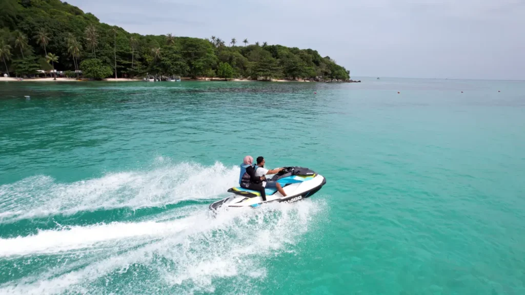 Pemandangan udara jet ski di laut jernih Karimunjawa dengan dua pengendara, berlatar pantai berpasir dan hutan hijau. Cocok untuk liburan pasangan